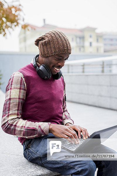 Lächelnder Mann mit Kopfhörer auf der Bank sitzend mit Laptop