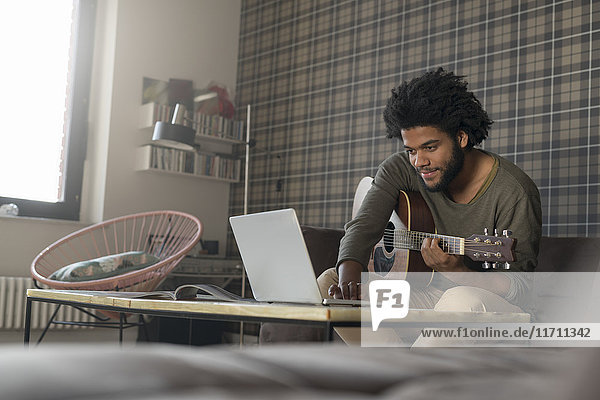 Mann sitzt im Wohnzimmer auf dem Sofa und spielt Gitarre vor dem Laptop.