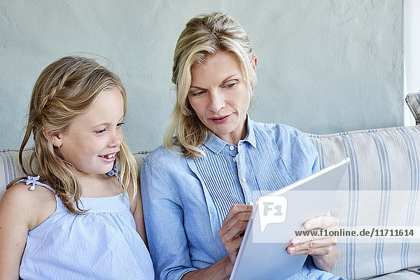 Mother and little daughter sitting together on couch looking at tablet