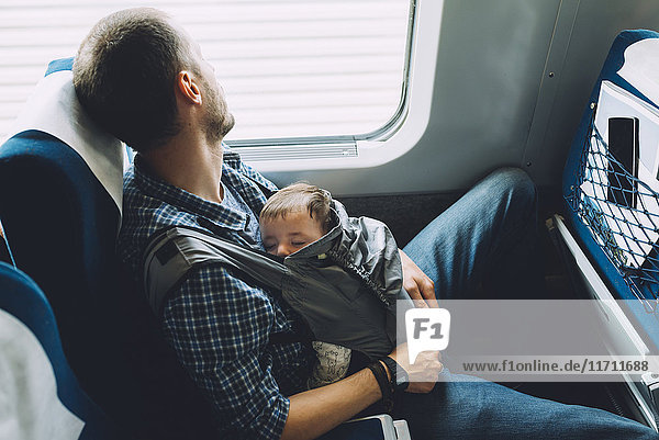 Vater im Zug  der sein schlafendes Baby in einem Tragesitz hält.