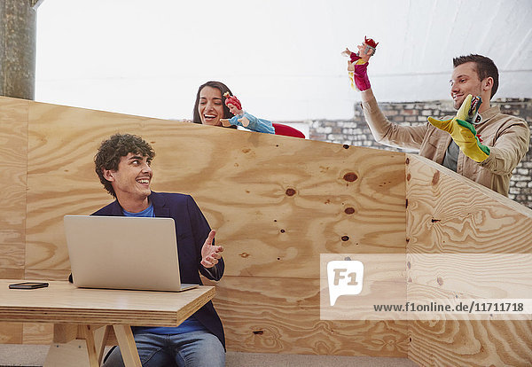 Junger Geschäftsmann arbeitet am Laptop mit Kollegen  die hinter einer Holzwand Handpuppen spielen.