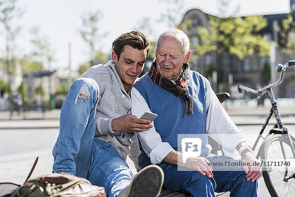 Senior man and adult grandson on a bench looking at cell phone
