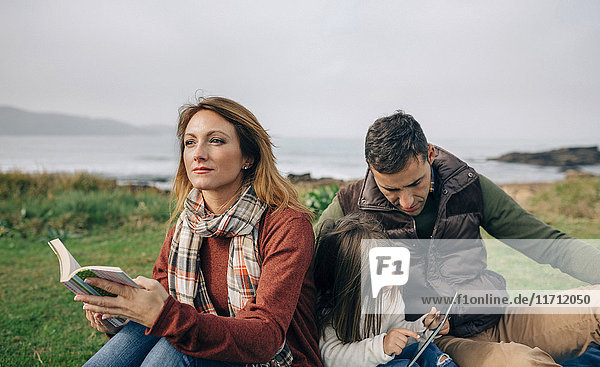 Relaxed woman with book looking aside while girl and man using tablet outdoors