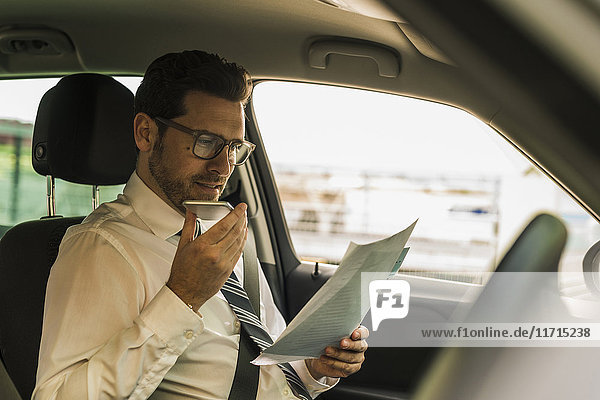 Successful businessman sitting in car reading files and using smartphone