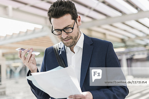 Businessman standing on parking level  reading documents  speaking notes on smartphone