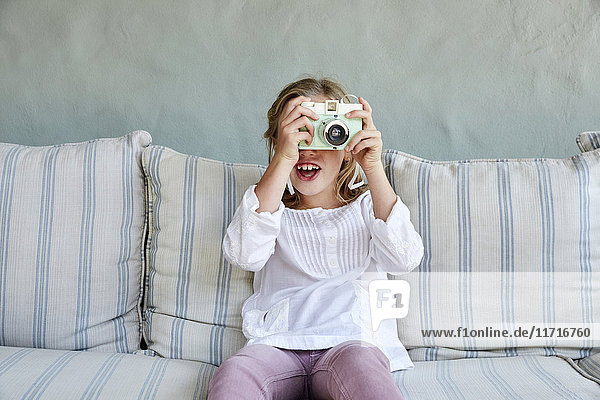 Portrait of smiling little girl sitting on couch taking picture with camera