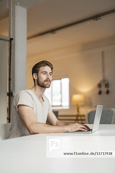 Porträt eines jungen Mannes  der auf einem Laptop in einem Loft arbeitet.