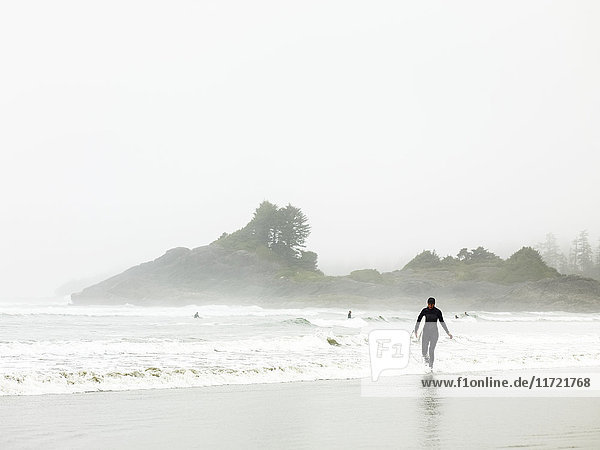 'A surfer in a wet suit walks along the water's edge  Cox Bay; Tofino  British Columbia  Canada'