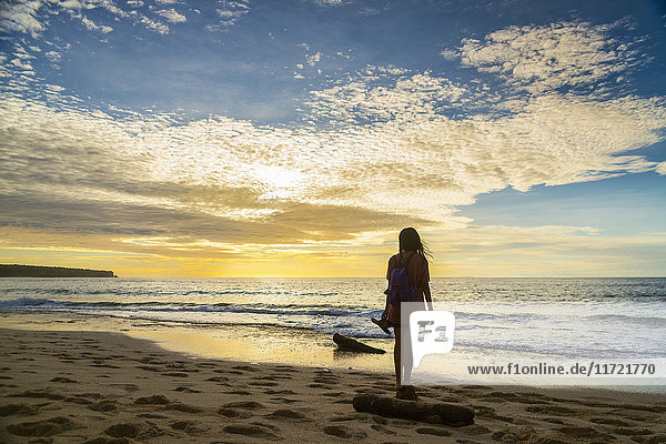 Eine Frau steht am Ufer und blickt bei Sonnenuntergang auf den Ozean  Dreamland Beach; Insel Bali  Indonesien'.