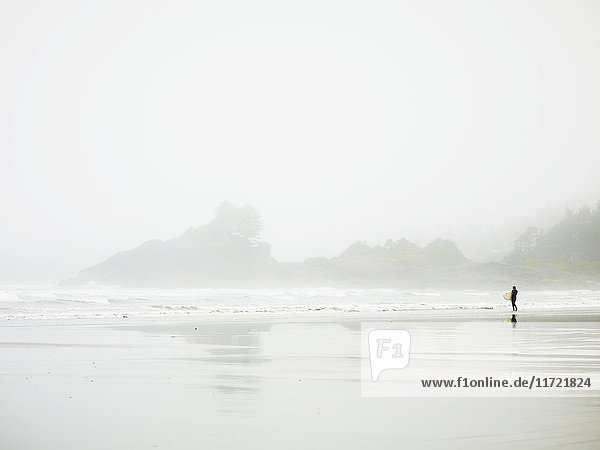 Ein einsamer Surfer steht am nassen Strand am Rande des Wassers  Cox Bay; Tofino  British Columbia  Kanada'.