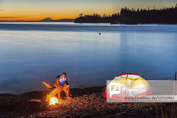 Ein Lagerfeuer am Strand bei Sonnenuntergang beleuchtet ein nahegelegenes Zelt. Ein Mann sitzt am Feuer und schaut auf sein Handy  während das ruhige Meerwasser das warme Licht im Hintergrund reflektiert; Hesketh Island  Homer  Alaska  Vereinigte Staaten von Amerika'.
