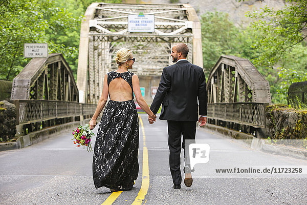 'A bride and groom holding hands and walking down a paved road; Oregon  United States of America'