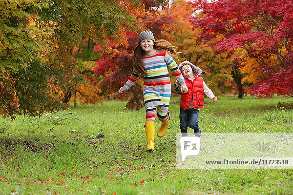 'A young boy and girl wearing rubber boots and colourful clothing run across a field holding hands with trees in bright autumn colours in the background; Oregon  United States of America'