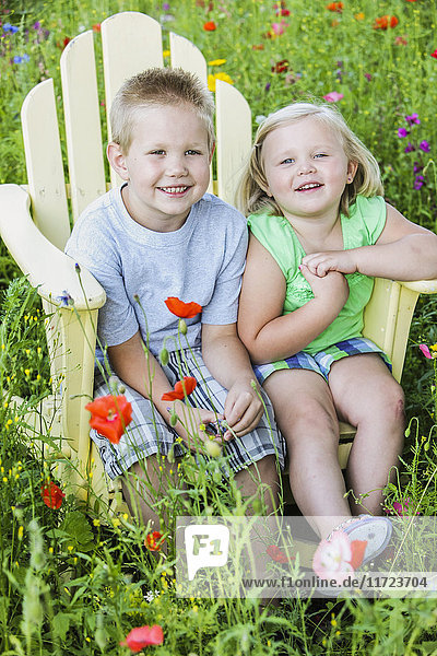 'Portrait of a young boy and girl sitting together in an adirondack chair surrounded by colourful wildflowers in a field; Oregon  United States of America'