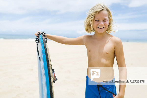 Junge (8-9) steht am Strand und hält ein Surfbrett