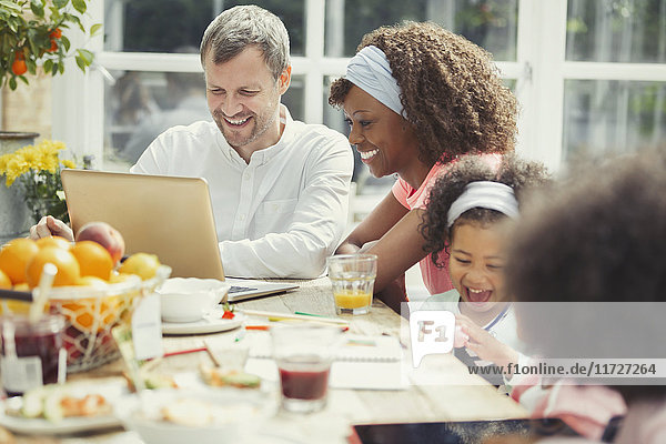 Lächelnde junge multiethnische Familie mit Laptop und Frühstück am Tisch