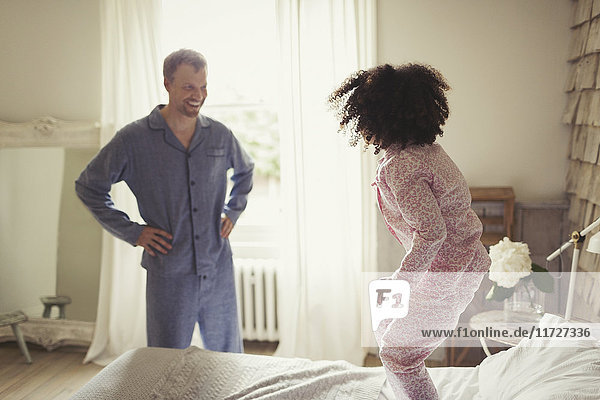 Ein multiethnischer Vater im Schlafanzug  der die Tochter beim Springen auf dem Bett beobachtet.