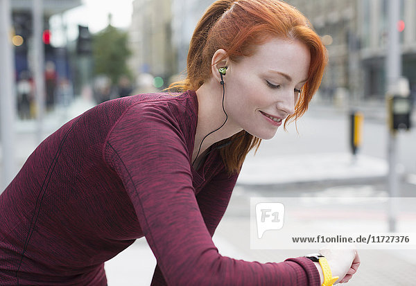 Lächelnde Läuferin mit roten Haaren und Kopfhörern  die auf intelligente Uhr achtet