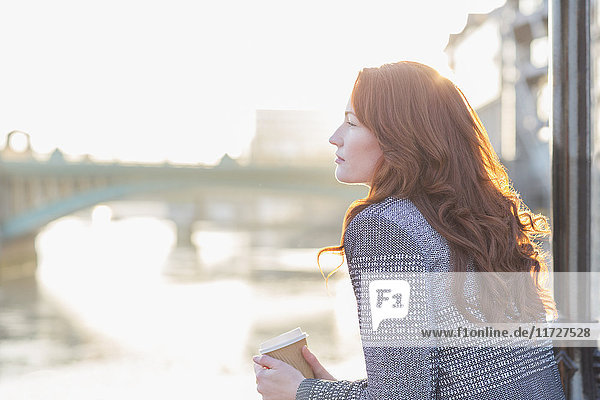 Gelassene Geschäftsfrau mit roten Haaren trinkt Kaffee an der städtischen Uferpromenade