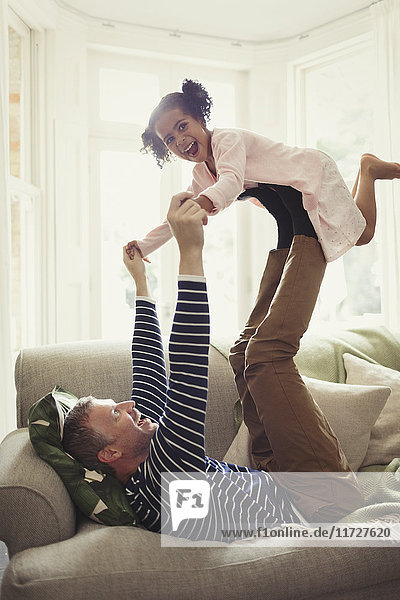 Playful multi-ethnic father balancing daughter on legs overhead on sofa