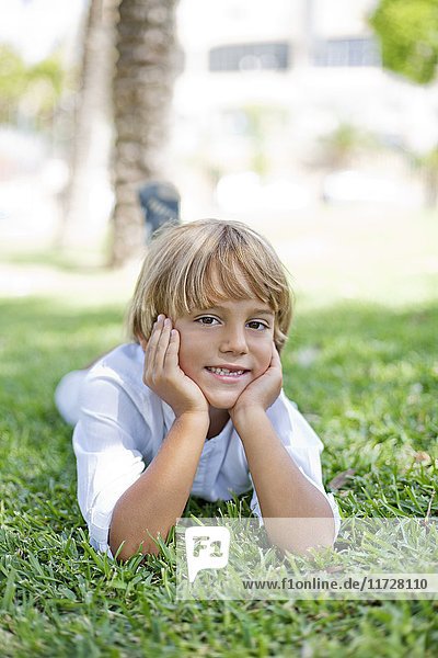 Süßer blonder Junge liegt auf Gras im Park und lächelt vor der Kamera.