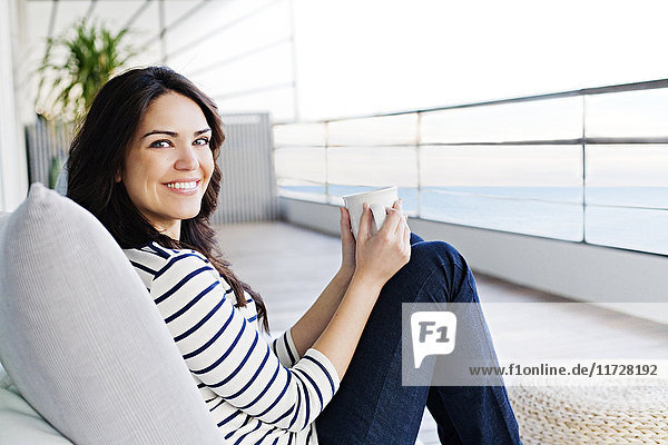 Schöne Frau genießt einen warmen Drink auf der Terrasse und lächelt vor der Kamera.