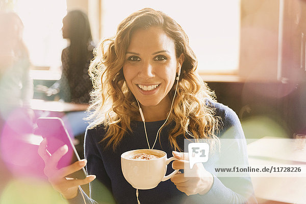 Porträt einer lächelnden Frau mit Kopfhörern  die Musik auf einem mp3-Player hört und Cappuccino in einem Café trinkt