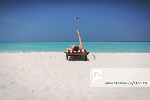 Frau liegt  sonnt sich und macht ein Selfie mit Selfie-Stick auf einem Liegestuhl am sonnigen tropischen Strand
