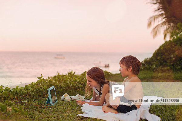 Junge und Mädchen Bruder und Schwester beobachten Video auf digitalen Tablet im Gras mit Blick auf das Meer