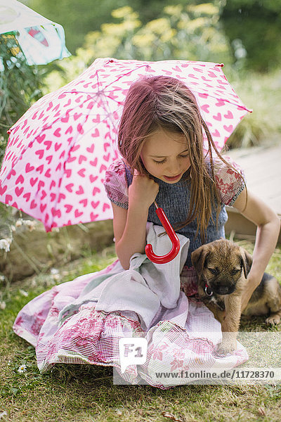Mädchen mit Hundewelpe hält herzförmigen Regenschirm im Gras