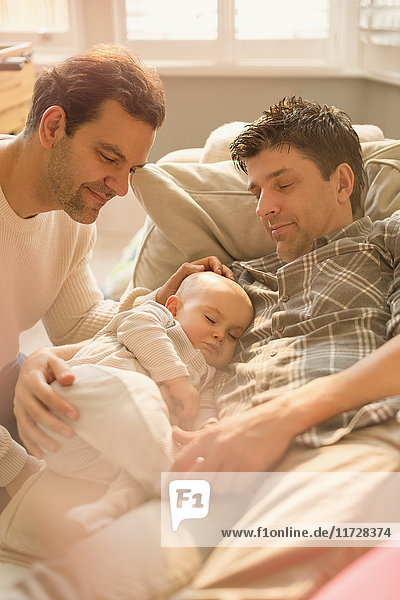 Male gay parents watching baby son sleeping on sofa