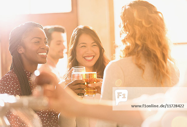 Lächelnde Freundinnen stoßen mit Biergläsern in einer sonnigen Bar an