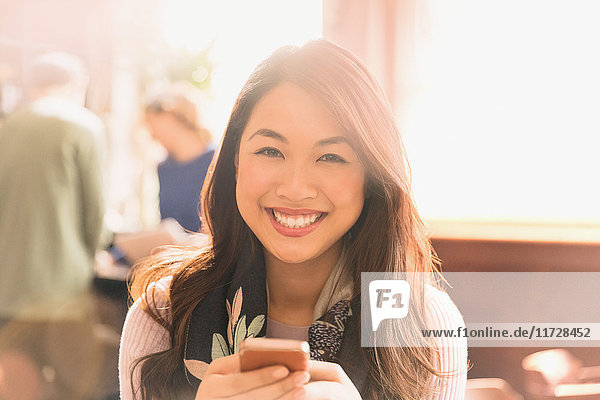 Porträt einer lächelnden chinesischen Frau  die in einem Café eine SMS mit ihrem Handy schreibt
