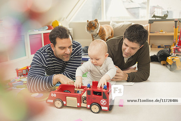 Male gay parents and baby son playing with fire engine toy in playroom