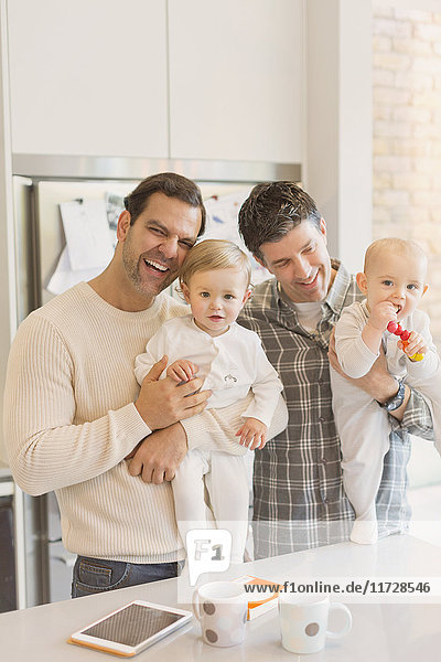 Portrait male gay parents holding baby sons in kitchen