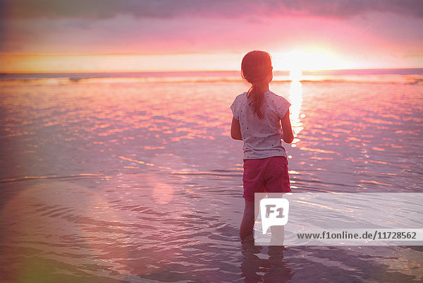 Nachdenkliches Mädchen watet im Meer am ruhigen Strand bei Sonnenuntergang