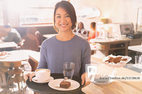 Porträt einer lächelnden Kellnerin  die ein Tablett mit Cappuccino  Brownie und Wasser in einem Cafe trägt