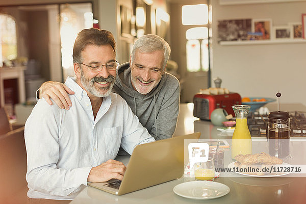 Male gay couple using laptop and eating breakfast at kitchen counter