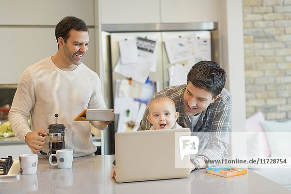 Male gay parents and baby son using laptop and digital tablet in kitchen