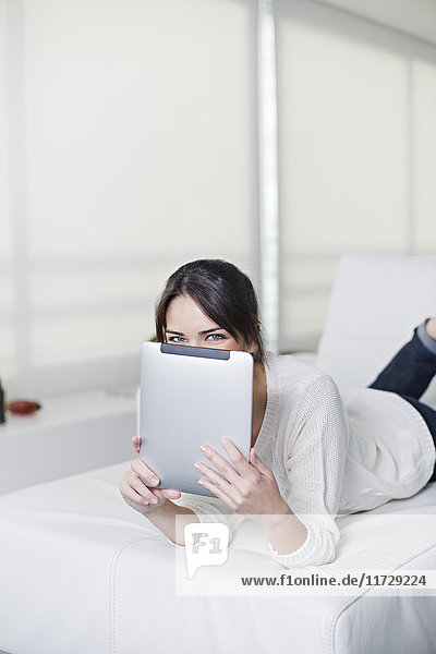 Verspielte brünette Frau auf einer Couch mit Tablette lächelnd vor der Kamera