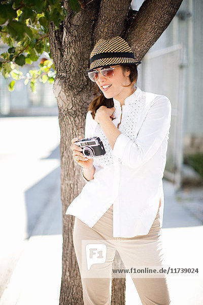 Mittelgroße erwachsene Frau mit Sonnenbrille und Hut  die eine alte Kamera hält und lächelt