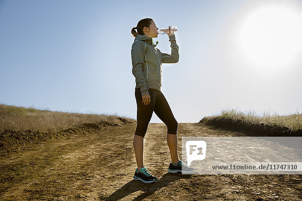 Silhouettierte Läuferin trinkt aus Wasserflasche auf Feldweg