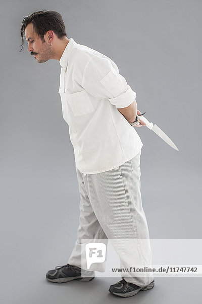 Seitenansicht Studioporträt eines männlichen Kochs mittlerer Größe mit Messer hinter dem Rücken