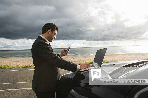 Geschäftsmann simst auf Smartphone  während er mit seinem Laptop auf dem Parkplatz an der Küste unterwegs ist