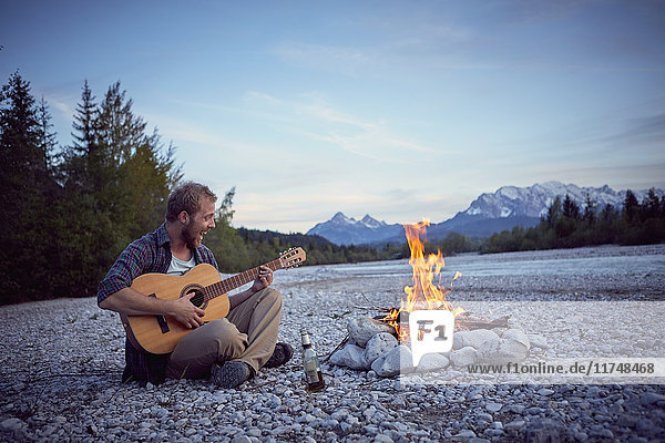 Junger Mann sitzt am Lagerfeuer  spielt Gitarre  singt  Wallgau  Bayern  Deutschland