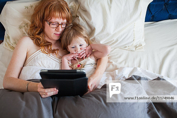 Mutter und Tochter liegen im Bett und schauen auf das digitale Tablett