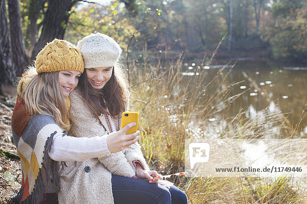 Teenagermädchen in Strickwaren sitzen am Fluss und benutzen ein Smartphone  um sich selbst zum Lächeln zu bringen