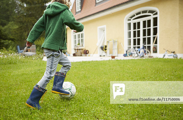 Rear view of boy wearing wellingtons playing football in the garden