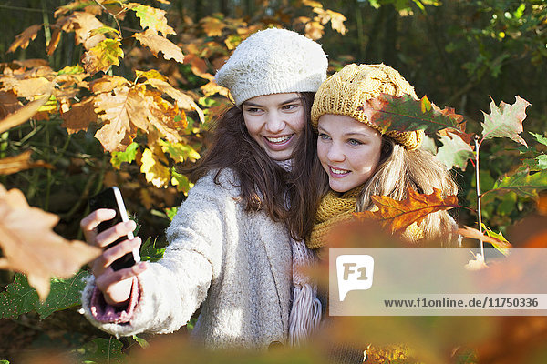 Schrägansicht von Teenager-Mädchen im Herbstlaub mit Smartphone  um Selfie zum Lächeln zu bringen