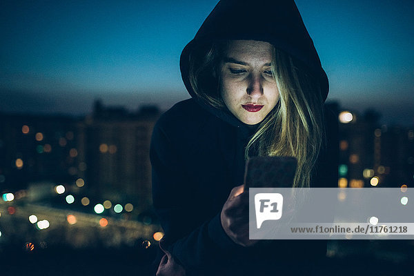 Junge Frau sitzt nachts auf dem Dach  benutzt Smartphone  erleuchtetes Gesicht